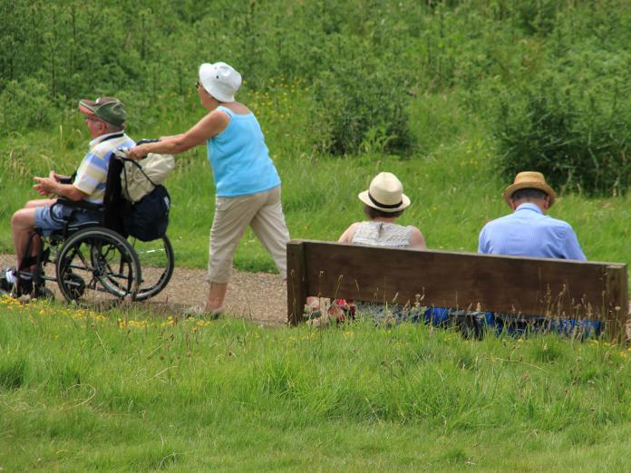 En mand og en kvinde sidder på en bænk med ryggen til. Ved siden af er en ældre kvinde ved at skubbe en person i kørestol op ad en bakke. 