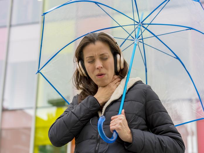 En kvinde står under en paraply og tager sig til halsen, som om hun har ondt i halsen. 